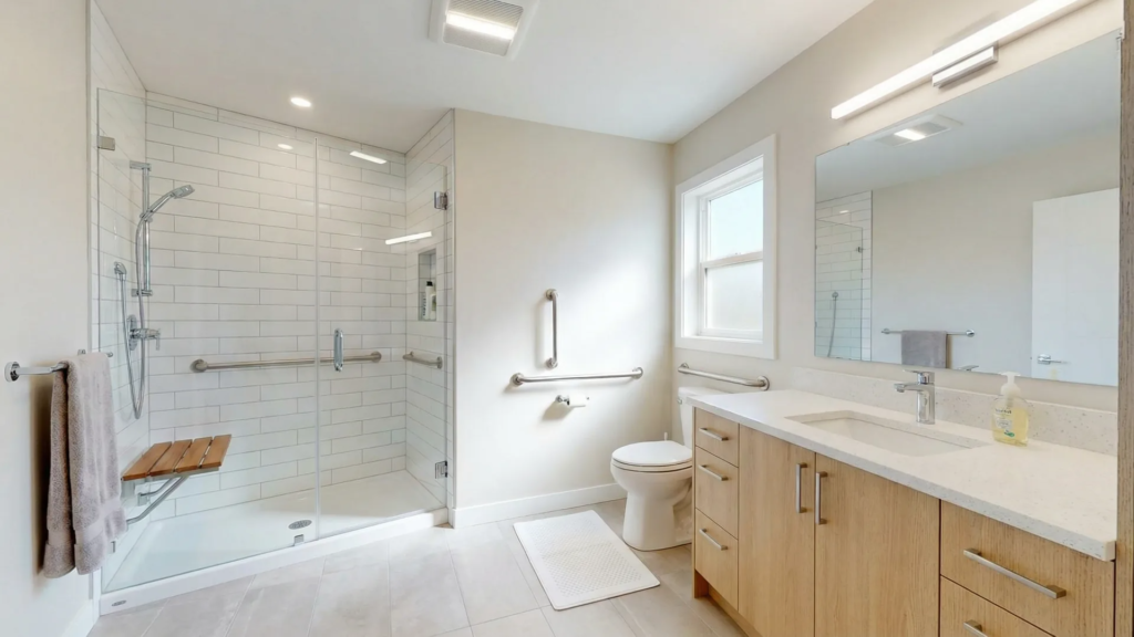 Senior-friendly bathroom in a Victoria BC home featuring chrome grab bars beside the toilet and shower, a non-slip bath mat, and a fold-down shower bench for fall prevention